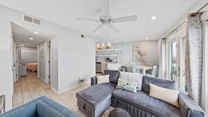 A modern living room with a grey sectional sofa, a blue armchair, and a round ottoman. There is a ceiling fan, a dining area with a table and chairs, and a kitchen in the background.