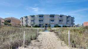 A beachfront apartment building with multiple balconies, a sandy pathway leading to the entrance, and a clear blue sky.