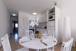 A dining area with a white table set for four, surrounded by white chairs. A white cabinet with a microwave and a small refrigerator is visible in the background. - Castle Harbour 32 - One Bed (Los Cristianos)