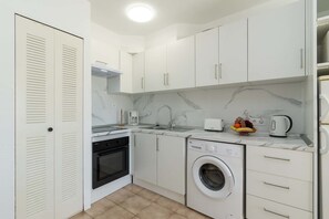 A compact kitchen with white cabinets, a built-in oven, a washing machine, and a coffee maker. - Castle Harbour 32 - One Bed (Los Cristianos)