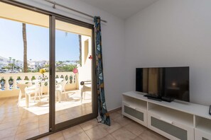 Habitación con puerta corrediza de vidrio que da a un espacio exterior con una mesa y sillas, un televisor sobre un mueble blanco y una cortina estampada.