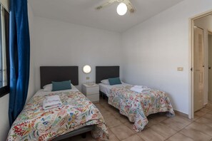 A bedroom with two beds, each with a floral bedspread, a bedside table with a lamp, and a window with blue curtains. - Castle Harbour 32 - One Bed (Los Cristianos)