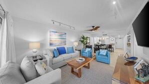A modern living room with a sofa, coffee table, and blue armchairs. There is a dining area with a table and chairs, and a kitchen in the background.