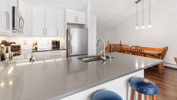 A modern kitchen with white cabinets, stainless steel appliances, a large island with stools, and a dining area with wooden chairs. - Quail Run Townhomes - Long Term Only - 30+ nights - Spacious, Garage, W/D, BBQ (Steamboat Springs)