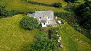 A stone house with a white door and windows, surrounded by a well-maintained lawn and dense shrubbery. - Kitts Cottage in Redruth (Redruth)