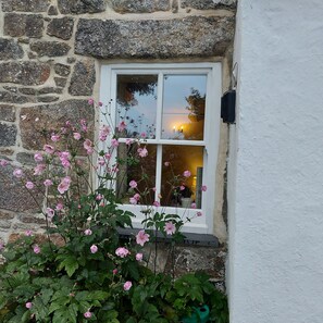 A stone wall with a white window and pink flowers in front. - Kitts Cottage in Redruth (Redruth)