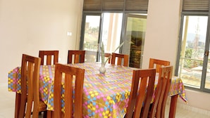 A dining room with a large table and wooden chairs, large windows, and a view of the outside. - OMX Hotel Remera (Kigali)