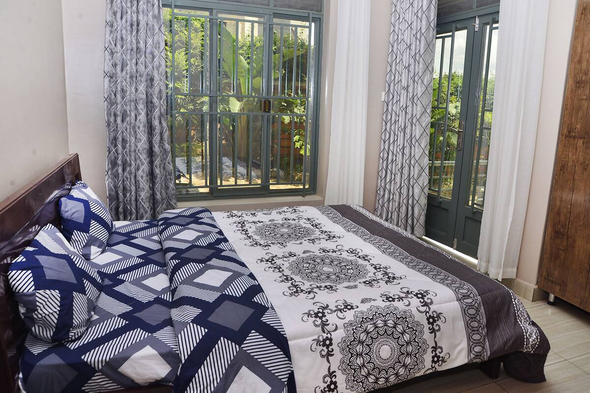 A bedroom with a bed, a window with a view of greenery, and a door with a metal gate.