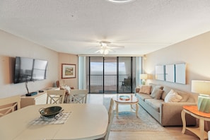 Sala de estar com televisão, ventilador de teto, mesa de jantar com um recipiente, sofá e varanda com vista.