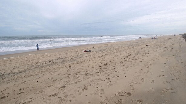 A beach with footprints leading to the water, a person fishing, and a cloudy sky.