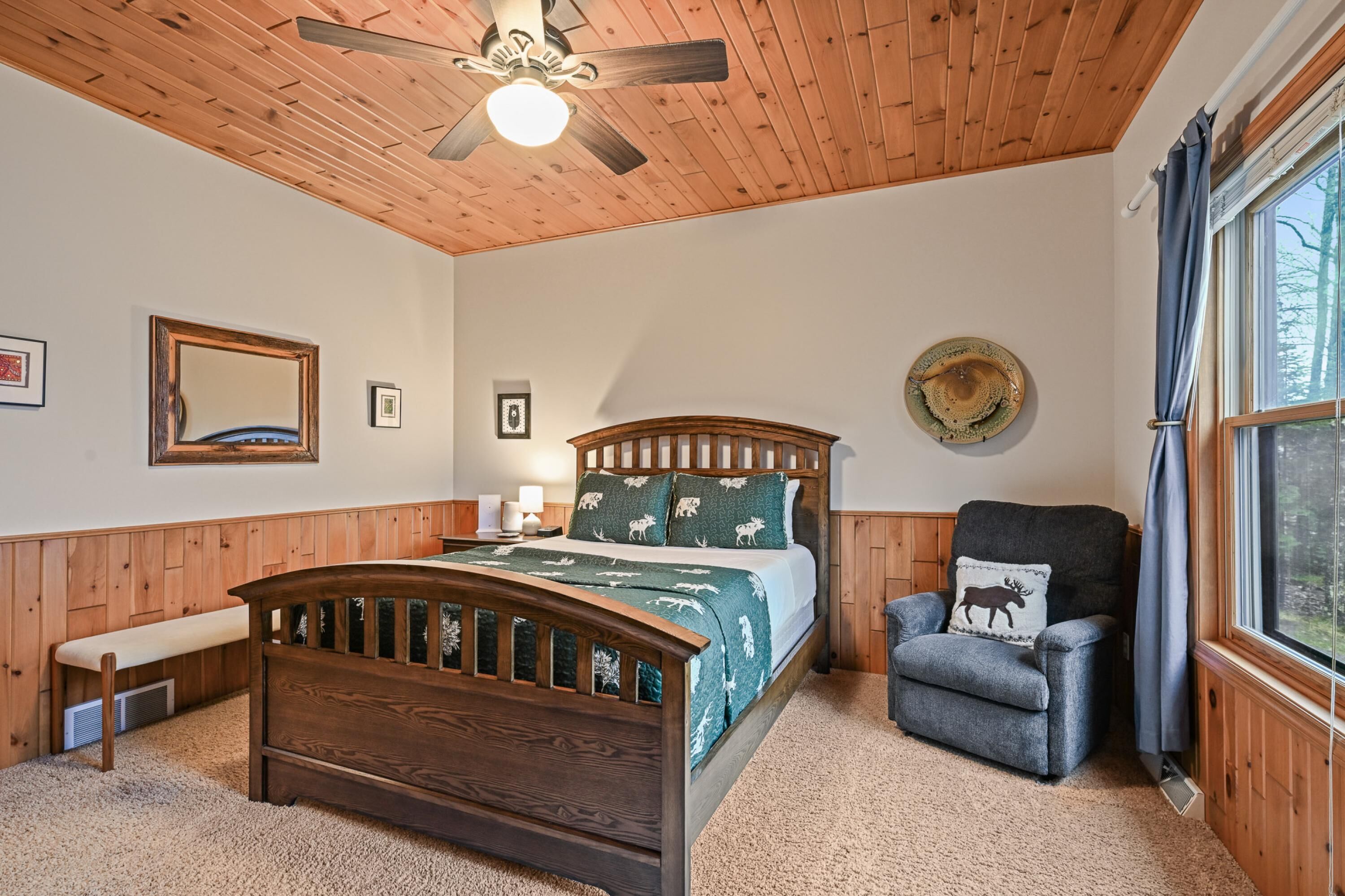 A bedroom with a wooden ceiling, a bed with a green and white bedding, a bench, a chair, and a window with curtains.
