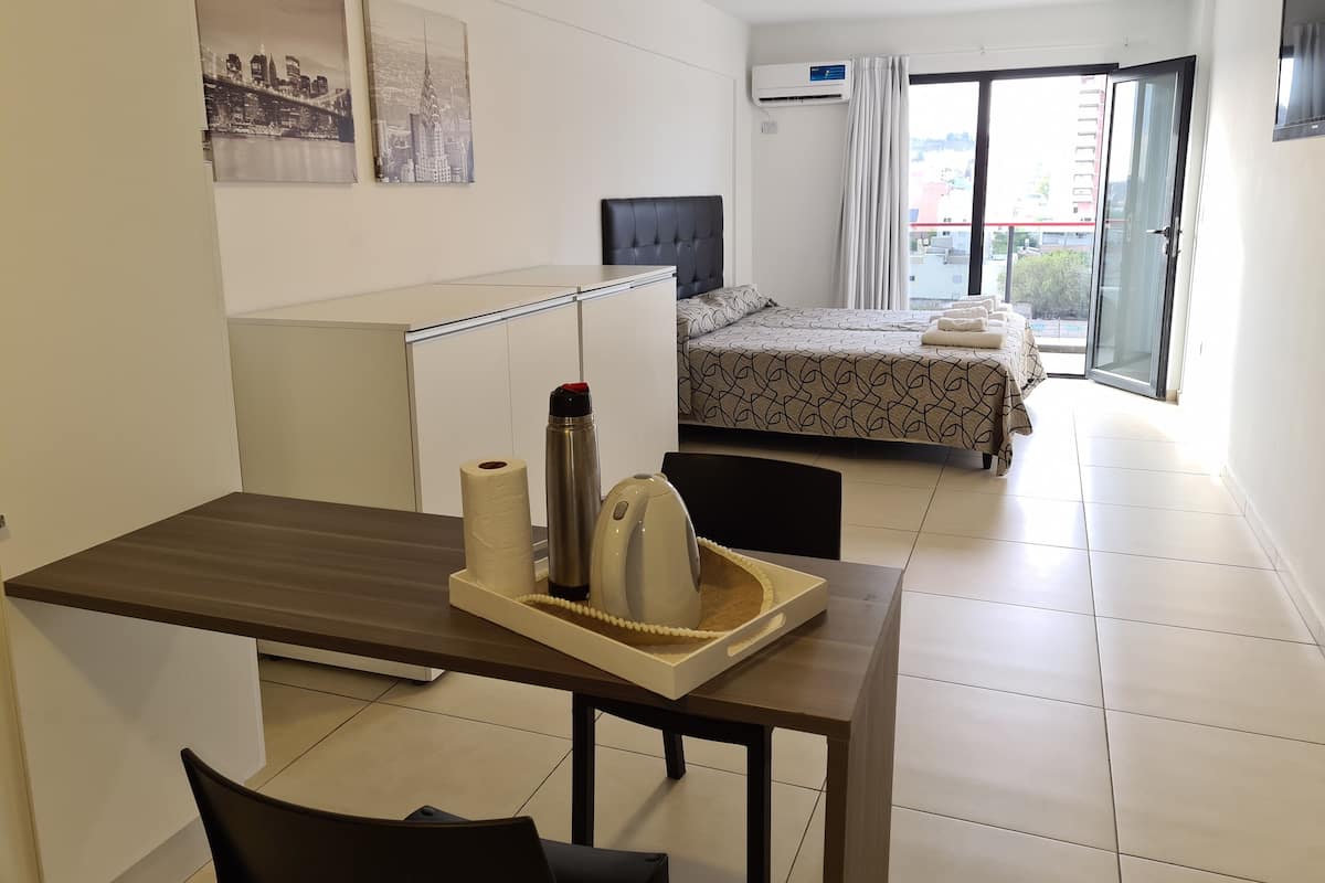 A hotel room with a bed, a desk, a chair, a table with a tray, and a window with curtains.