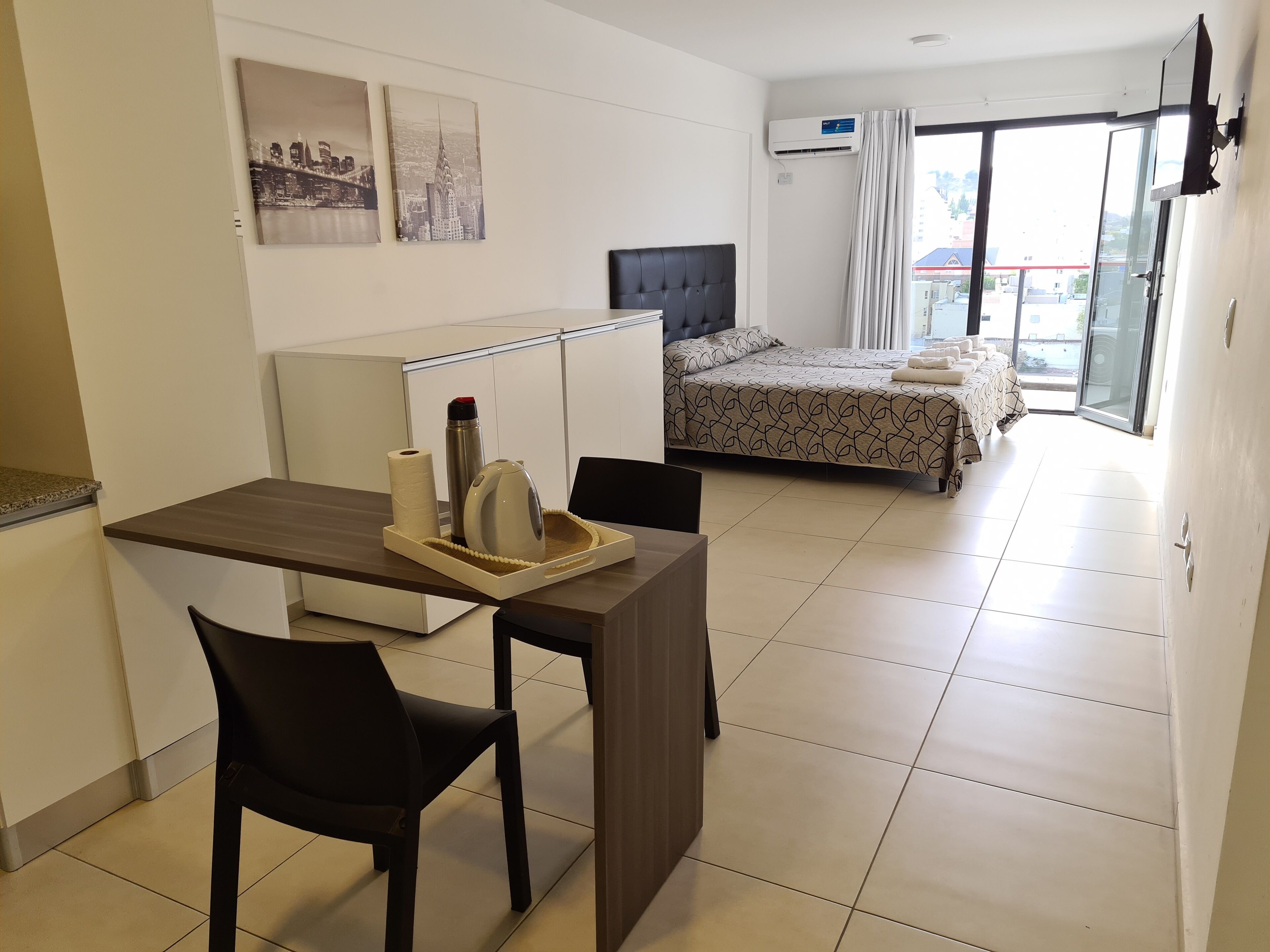 Quarto de hotel moderno com uma cama, uma escrivaninha com cadeira, uma mesinha de apoio com bandeja e uma janela com cortinas.