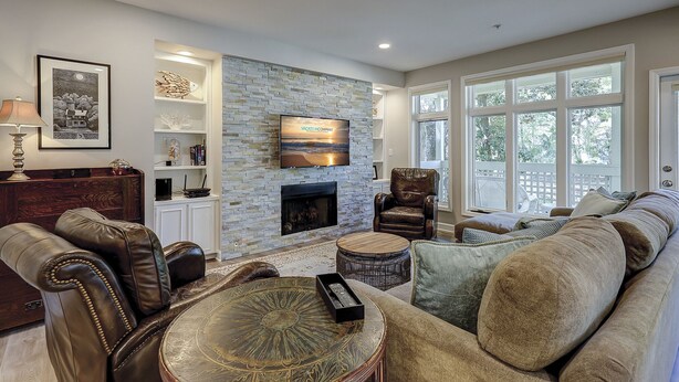 Una sala de estar con chimenea de piedra, mobiliario de cuero, televisor montado y ventanas amplias.