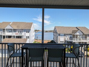 Un balcon avec des fauteuils en osier et une table, donnant sur un lac avec des bâtiments en arrière-plan.