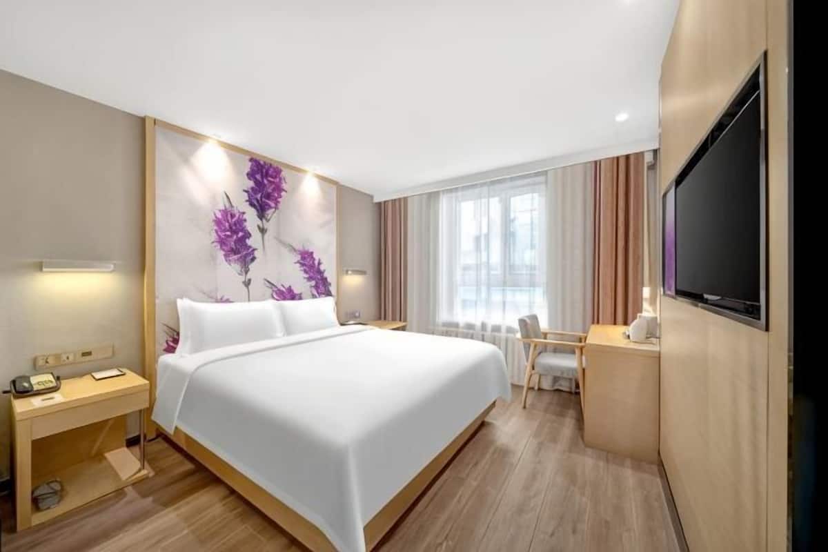 Habitación de hotel con cama, mesita de noche, escritorio, silla, televisión y un amplio ventanal con cortinas.