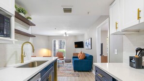 A modern kitchen with white countertops, dark cabinetry, and gold handles. There is a built-in microwave, a sink, and a window with blinds. A blue sofa and a small rug are in the living area, and a flat-screen TV is mounted on the wall. - Chic Condo Retreat in Central Buckhead Atlanta (Atlanta)