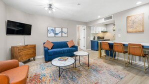 A modern living room with a blue sofa, two round tables, a TV, and a kitchen area with bar stools. - Chic Condo Retreat in Central Buckhead Atlanta (Atlanta)