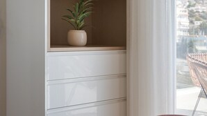 A bedroom with a bed, a white wardrobe, a shelf with a plant, and a view of a cityscape through a window. - Athenian Riviera Elegant Residence (Vari-Voula-Vouliagmeni)