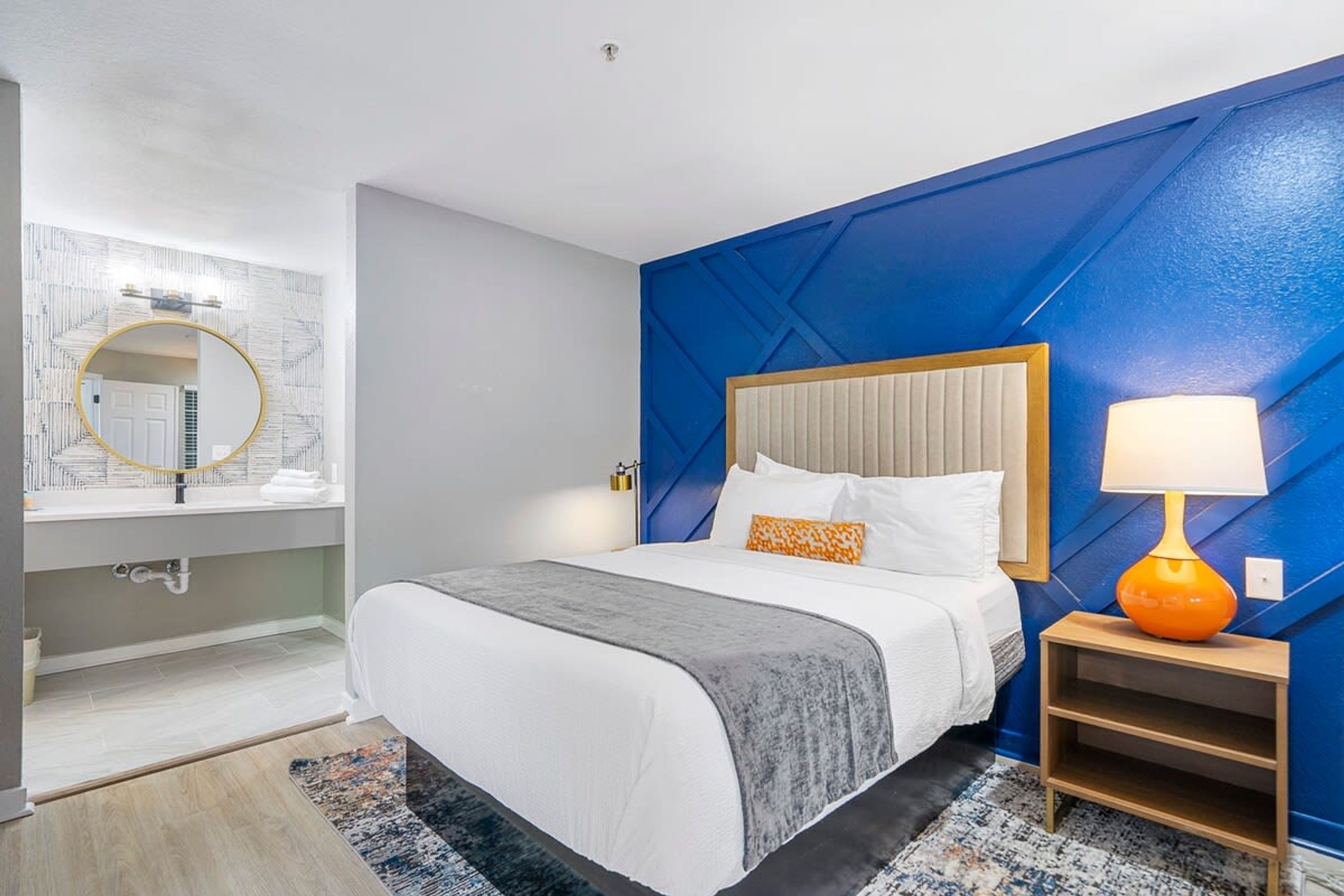 Ein modernes Hotelzimmer mit einem großen Bett, einem Nachttisch mit Lampe, einem Badezimmer mit Spiegel und Waschbecken sowie einer blauen Akzentwand.
