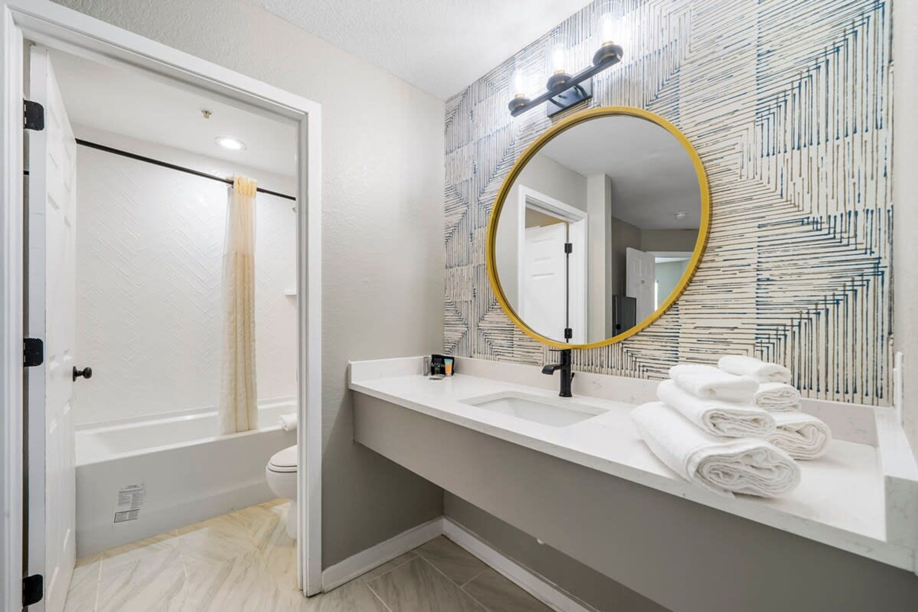 Un baño con tocador blanco, un espejo grande con marco dorado y un papel tapiz estampado.
