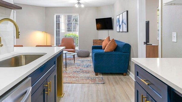 Une cuisine moderne dotée d’un îlot central, d’un coin repas avec une chaise, d’un canapé avec des coussins et d’un téléviseur fixé au mur.