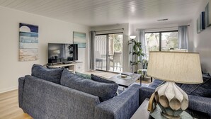 A modern living room with a flat-screen TV, grey sofas, a coffee table, and a large window with curtains.