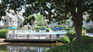 A pool area with a floating deck, surrounded by greenery and a building in the background.