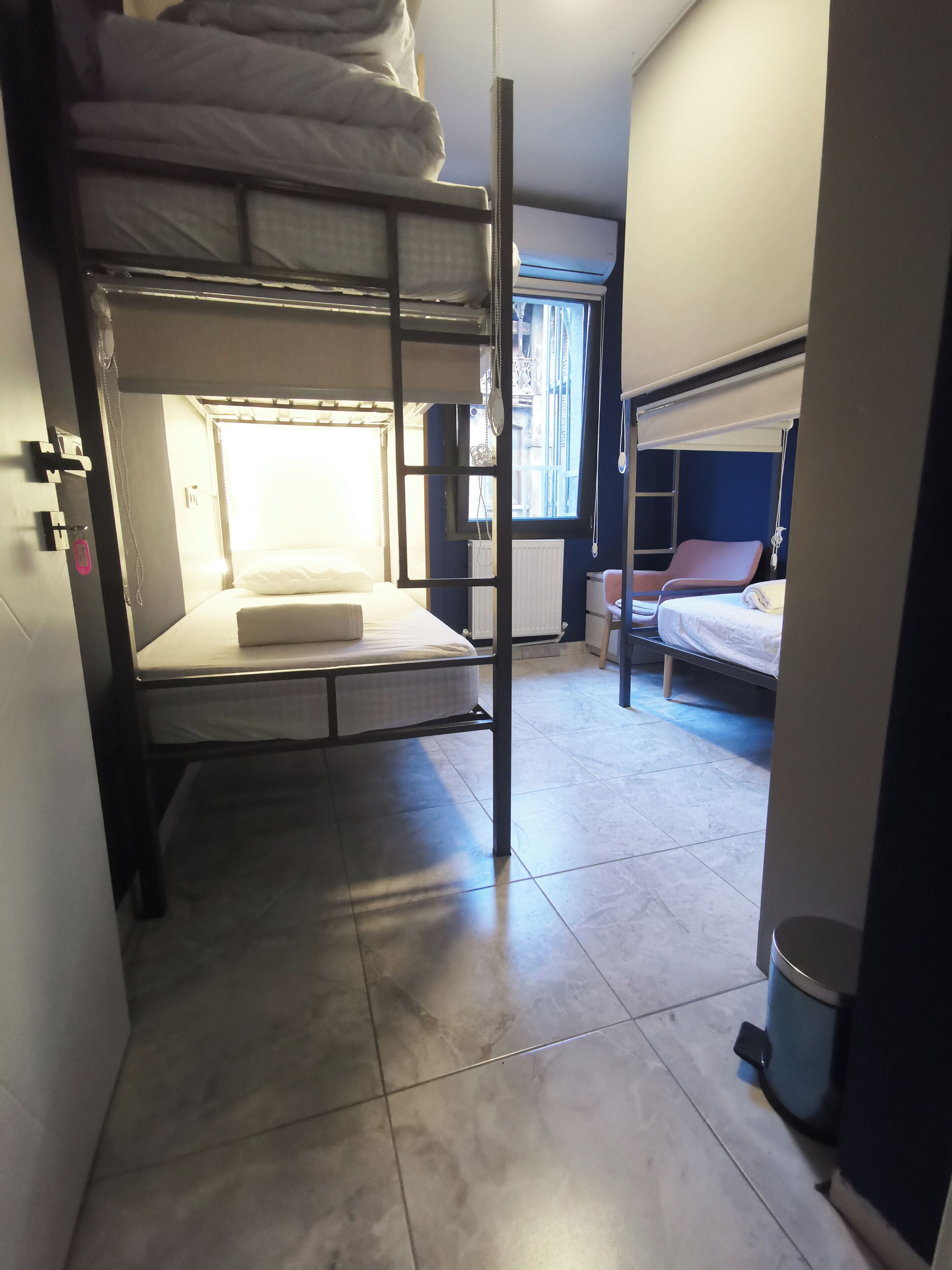 A dormitory room with bunk beds, a window with blinds, a radiator, and a trash bin.