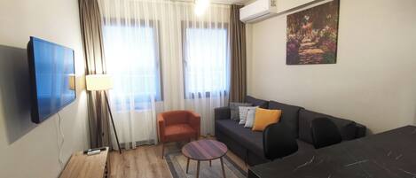 A modern living room with a flat-screen TV, a sofa with cushions, a coffee table, a dining table, and a painting on the wall.