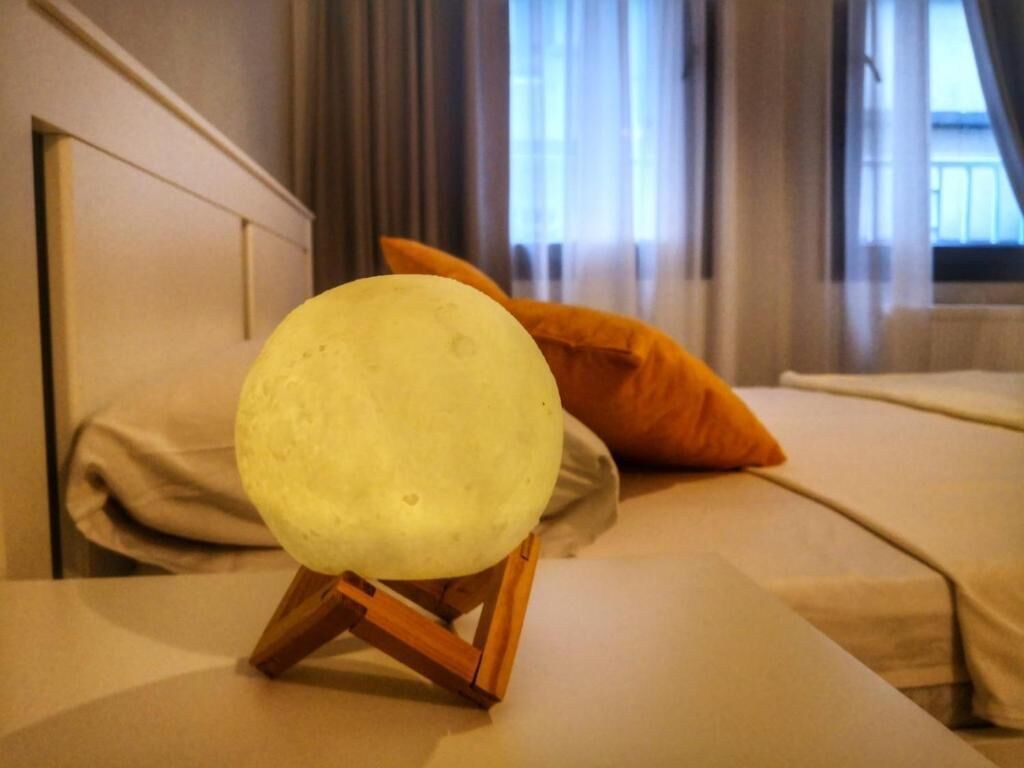 Un letto con un oggetto rotondo illuminato su un supporto in legno, un cuscino color beige e una finestra con tende sullo sfondo.