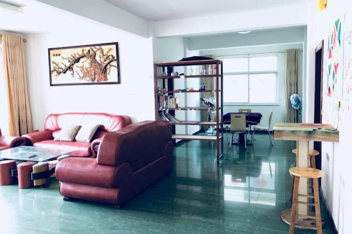 A modern living room with a red leather sofa, a glass coffee table, a wooden bookshelf, and a dining area with a table and chairs.