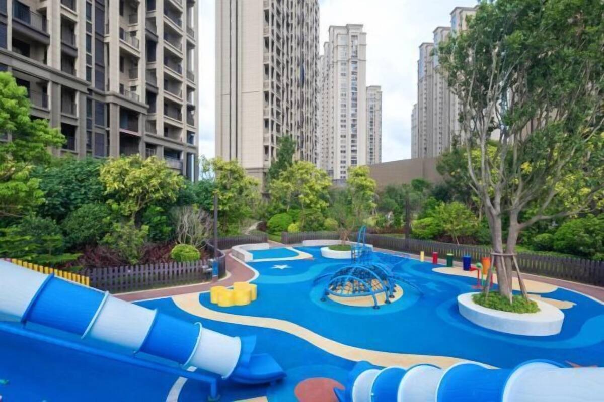 A playground with slides and climbing structures, surrounded by greenery and tall buildings.