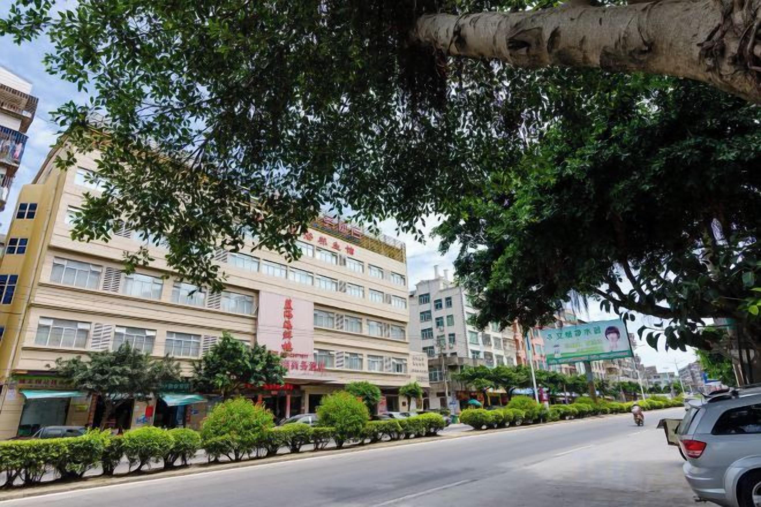 A multi-story hotel with a row of windows on the ground floor, a tree-lined street, and parked cars.