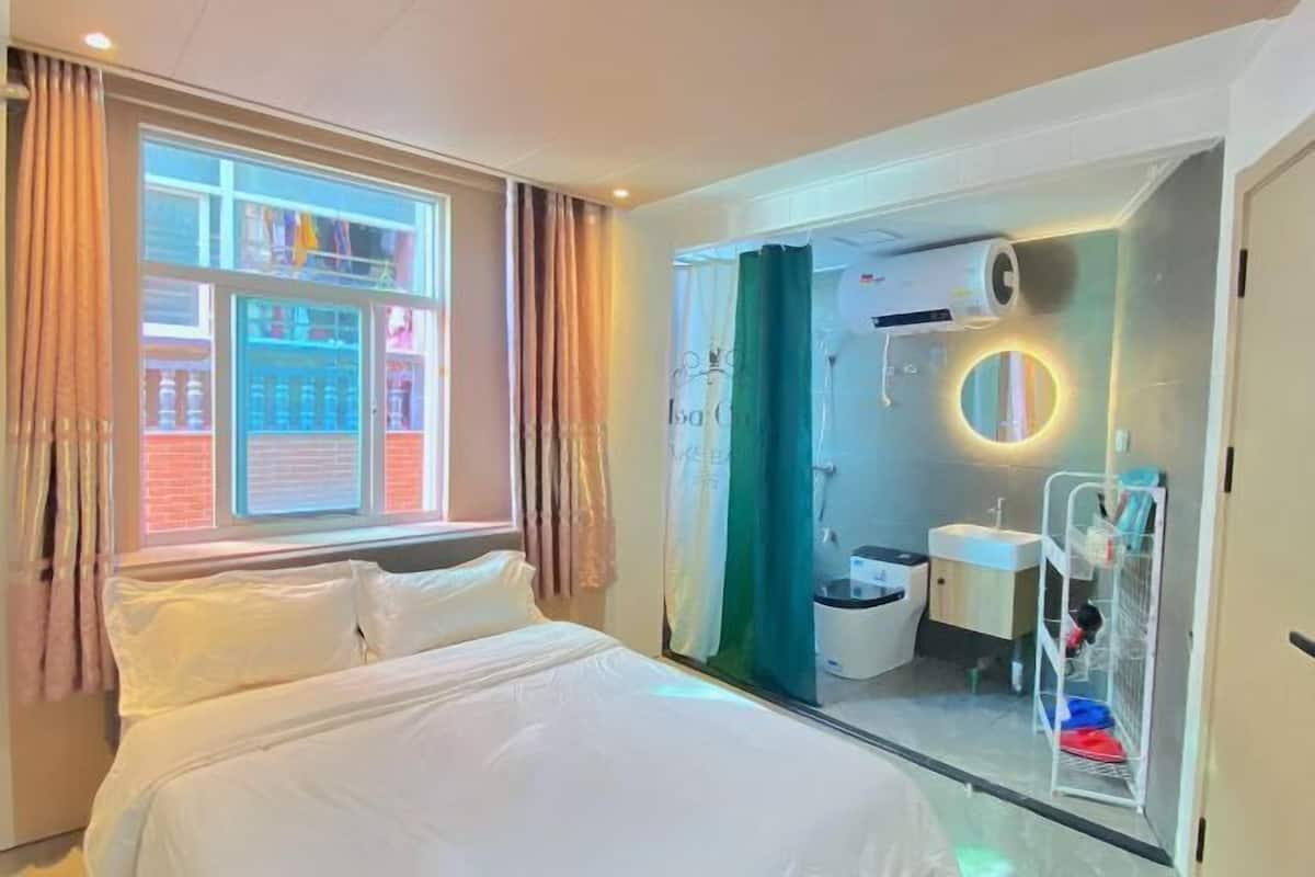 A hotel room with a bed, a window with curtains, a bathroom with a sink and mirror, and a shower area.
