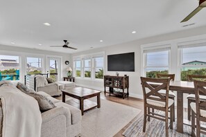A spacious living room with a large window, a ceiling fan, a flat-screen TV, and a dining area with wooden chairs and a table. - Oceanwatch Cottage | Spacious Retreat + Ocean Water Views! (Surf City)