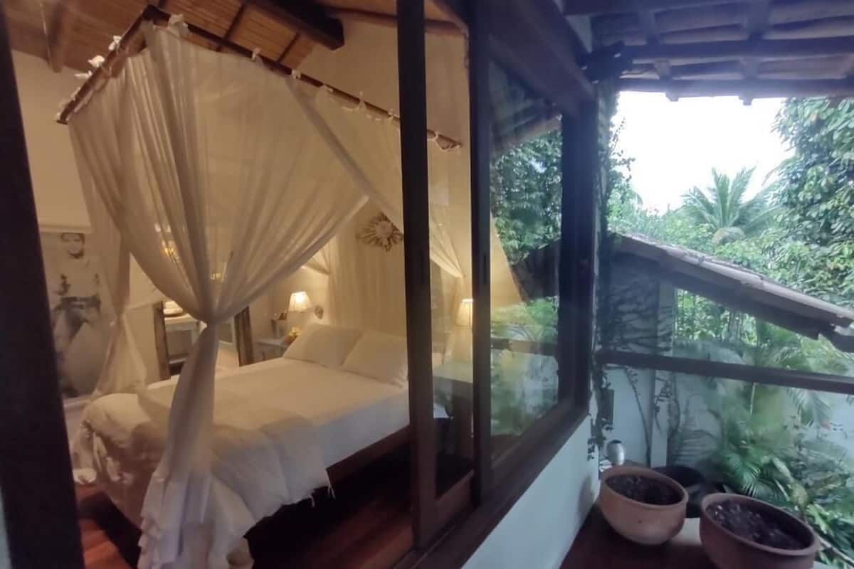 A bedroom with a canopy bed, a window with a view of greenery, and two potted plants on the windowsill.