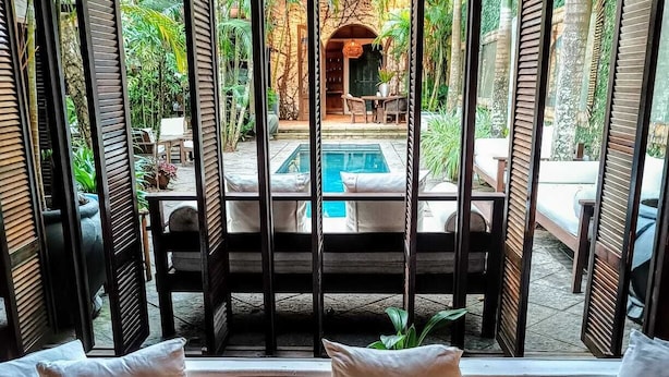 Une pièce dotée d’une grande fenêtre donnant sur une piscine et un jardin. On y trouve un canapé avec des coussins et une table avec des chaises.
