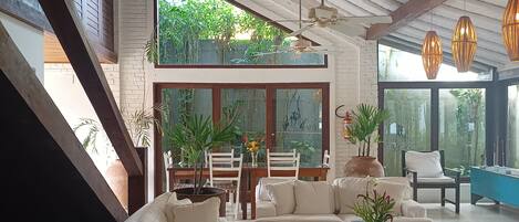 Amplia sala de estar con un sofá blanco grande, una mesa de centro de madera y una mesa de comedor con sillas. Hay plantas en macetas y vistas a zonas verdes al exterior.