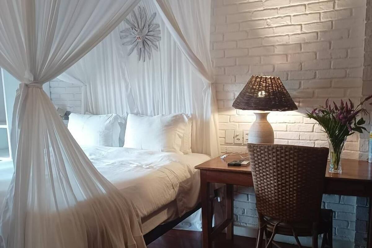 A bedroom with a canopy bed, a bedside table, a lamp, and a wicker chair.