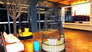 Un área de descanso moderna con techo de madera, un ventanal con vistas a la ciudad, una estructura de asientos de metal negro y un banco de madera con cojines.
