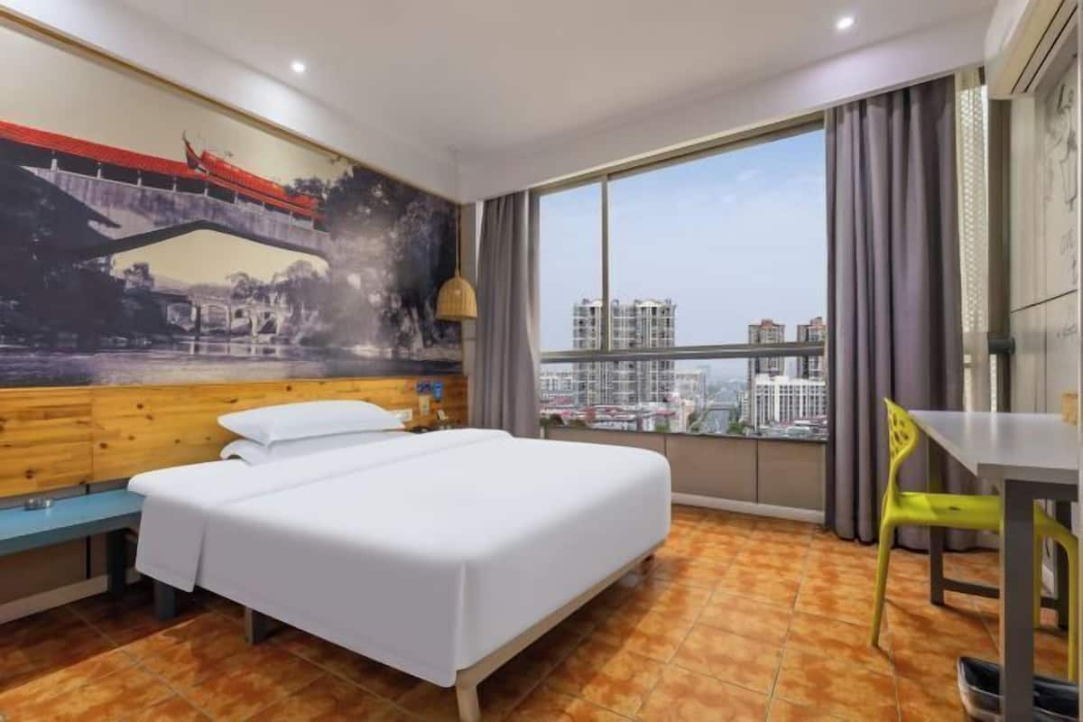 Habitación de hotel con cama, banca, mesa de comedor, silla, ventana grande con vista a la ciudad y un mural en la pared.
