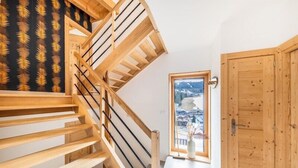 Eine Holztreppe mit einem Teppich in Schwarz mit Blattmuster, eine Holztür und ein Blick auf eine verschneite Landschaft durch ein Fenster.