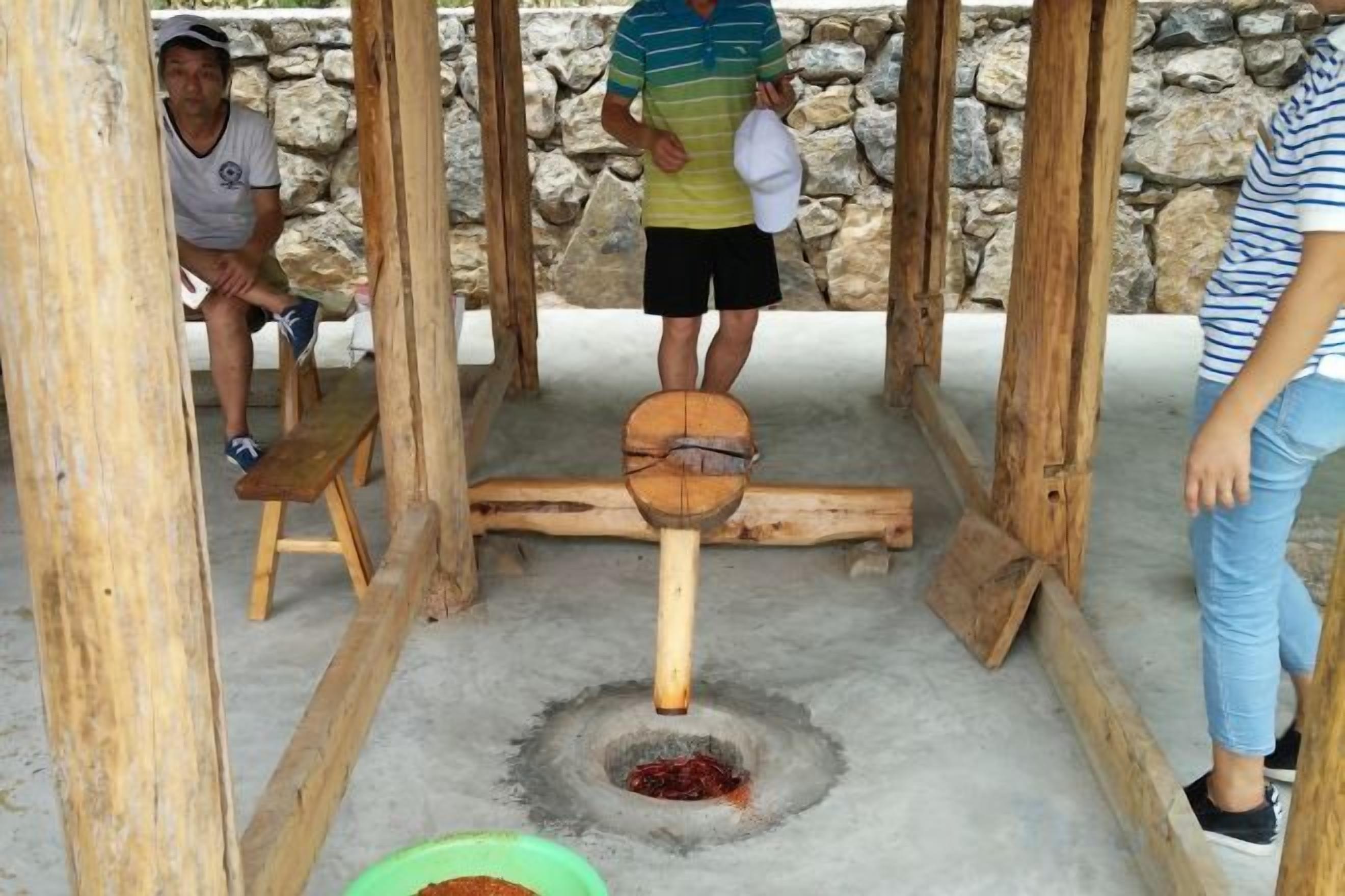 A rustic outdoor setting with a wooden structure, a stone wall, and a person operating a manual grinding device.