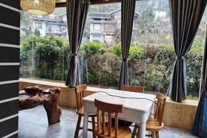 Ein Essbereich mit Tisch und Stühlen, einer Holzbank sowie Blick auf Grünflächen und Gebäude durch große Fenster.