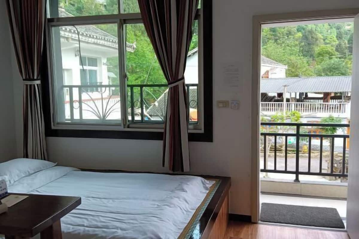 A bedroom with a bed, a window with curtains, a balcony with a railing, and a view of greenery outside.