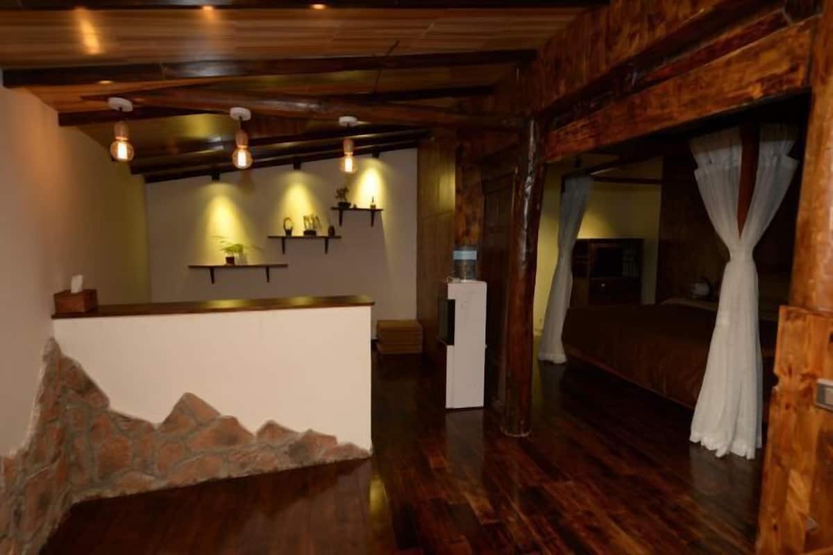 A room with a stone fireplace, wooden ceiling beams, a bed with white curtains, and shelves with decorative items.