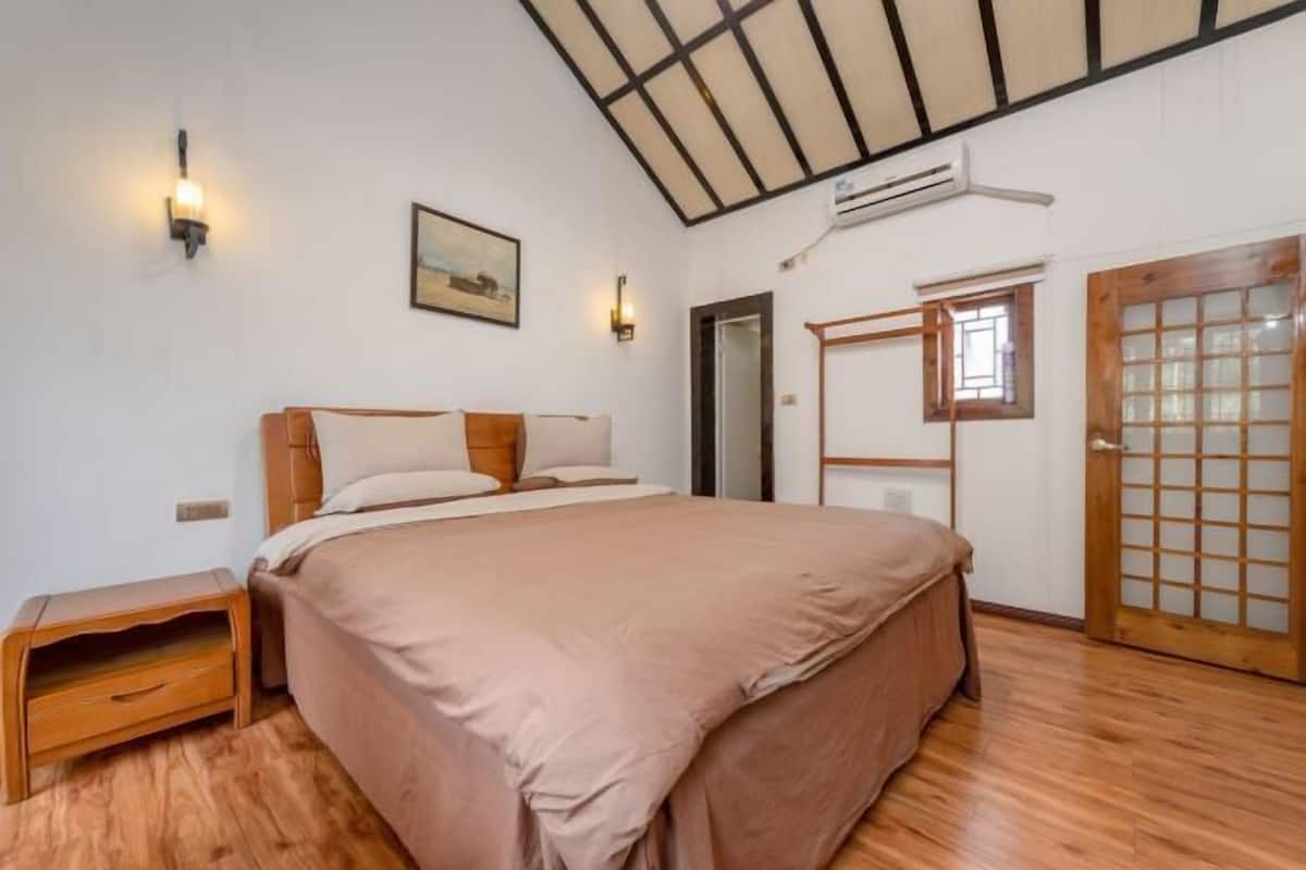 A bedroom with a large bed, wooden headboard, a nightstand, a wall-mounted light, a painting, and a sliding door with a grid pattern.