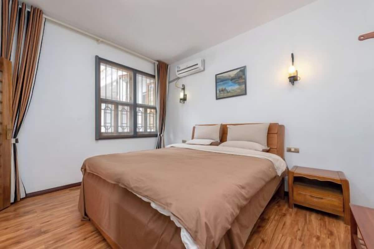 A bedroom with a bed, wooden nightstand, wall-mounted air conditioning unit, and a window with curtains.