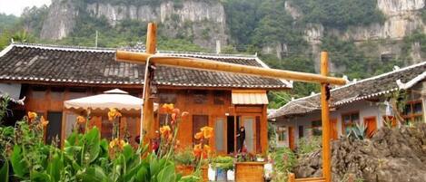 A traditional wooden house with a tiled roof, surrounded by lush greenery and a rocky cliff in the background.